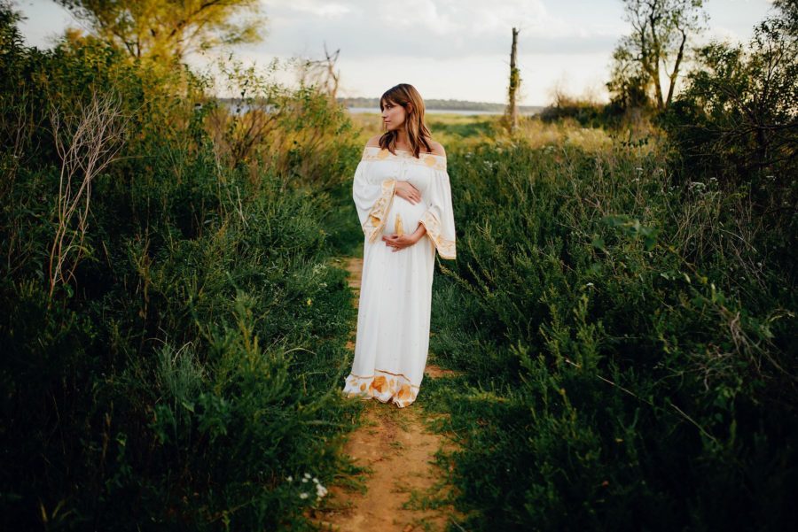 Pregnant woman holding belly wearing long white dress outside, Beyond the Wanderlust Daily Fan Favorites 