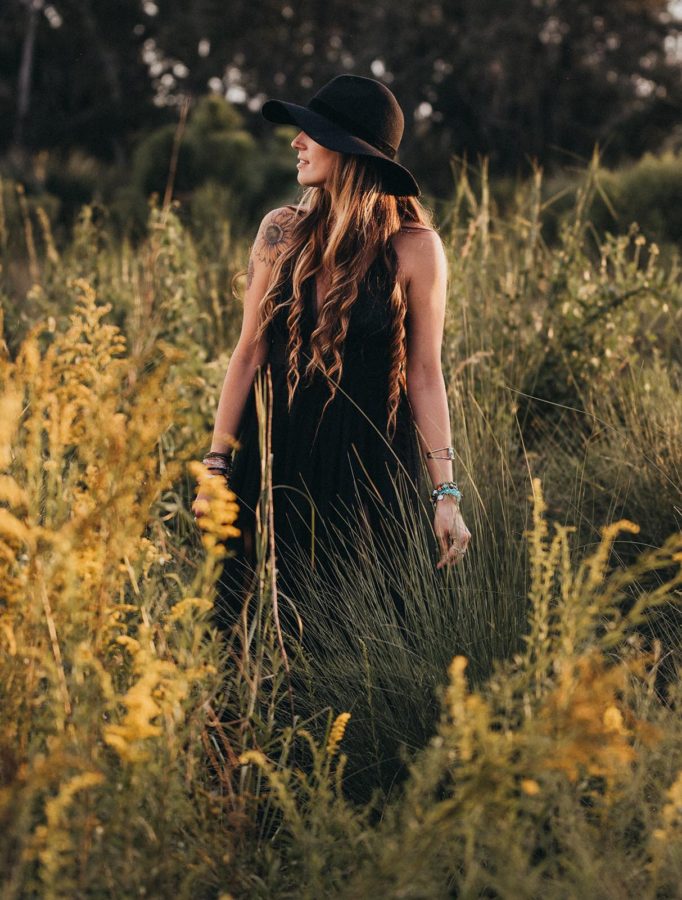 Woman with long hair standing in long grasses with black dress and hat, Beyond the Wanderlust Daily Fan Favorites