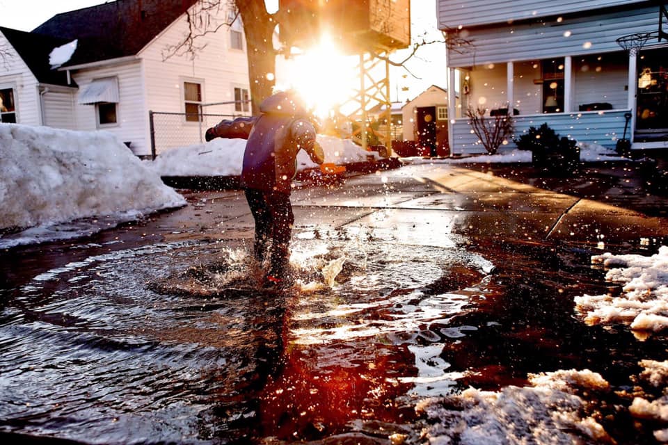 spring pictures of kids in snowy states, young boy jumping in snow puddles, sun flare, golden hour