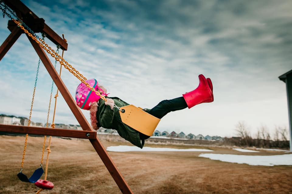 spring pictures of kids, little girl swinging high on swing set, little girl wearing pink rain boots, blue and cloudy sky